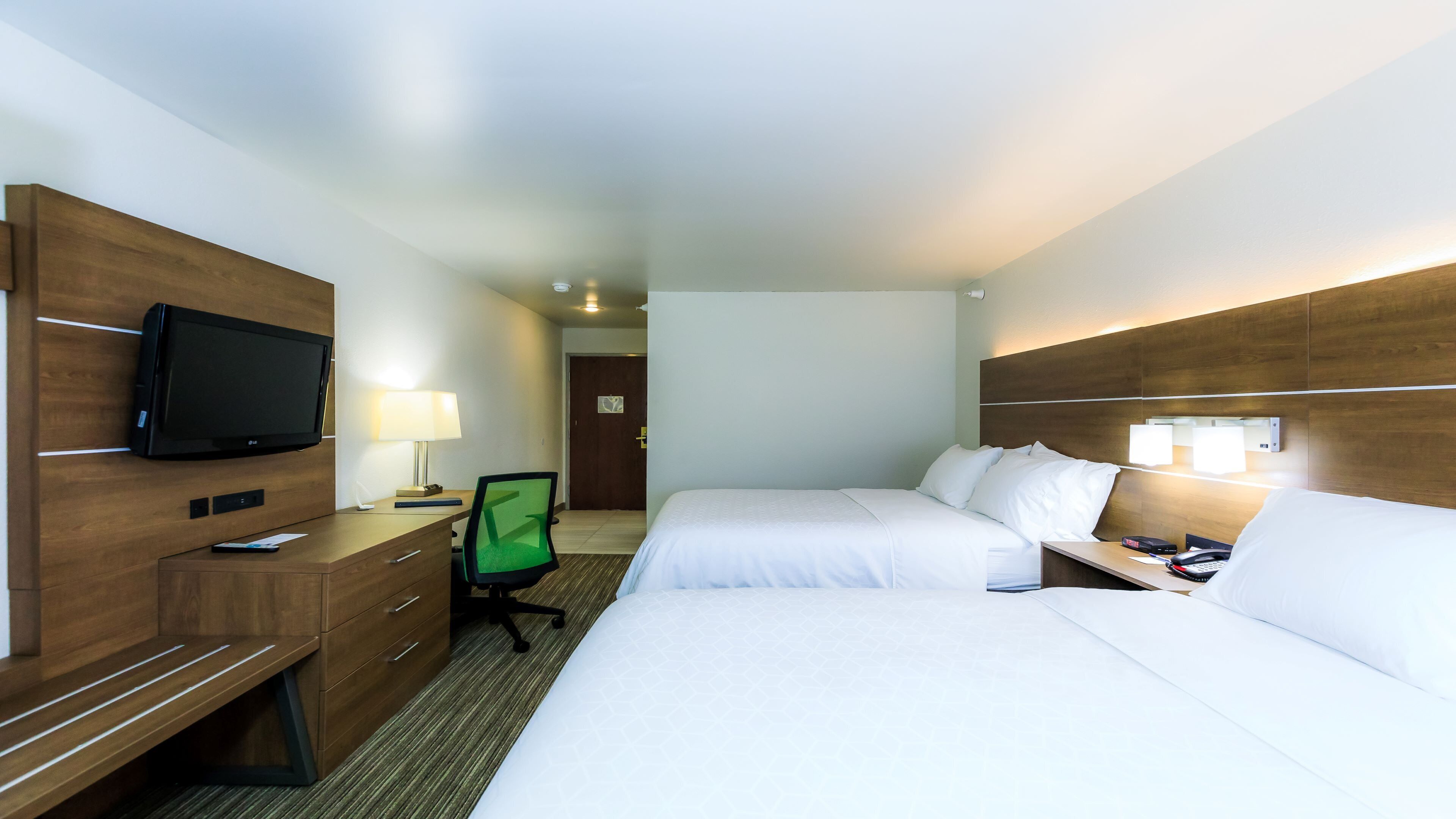 A hotel room with a bed, a desk with a flat-screen TV, a chair, and a bedside table with a lamp.