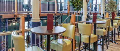Un restaurante con mesas de madera altas, sillas amarillas, una escalera y ventanas grandes.
