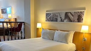 A hotel room with a large bed, two bedside lamps, a wooden headboard, a small wooden nightstand, and a red chair in the background.