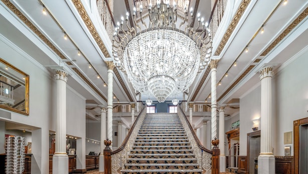A grand staircase with ornate railings and a large chandelier, leading to a high-ceilinged lobby with columns and a patterned floor.