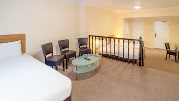 A hotel room with a bed, a glass coffee table, and four chairs. There is a staircase with a railing leading to another room.
