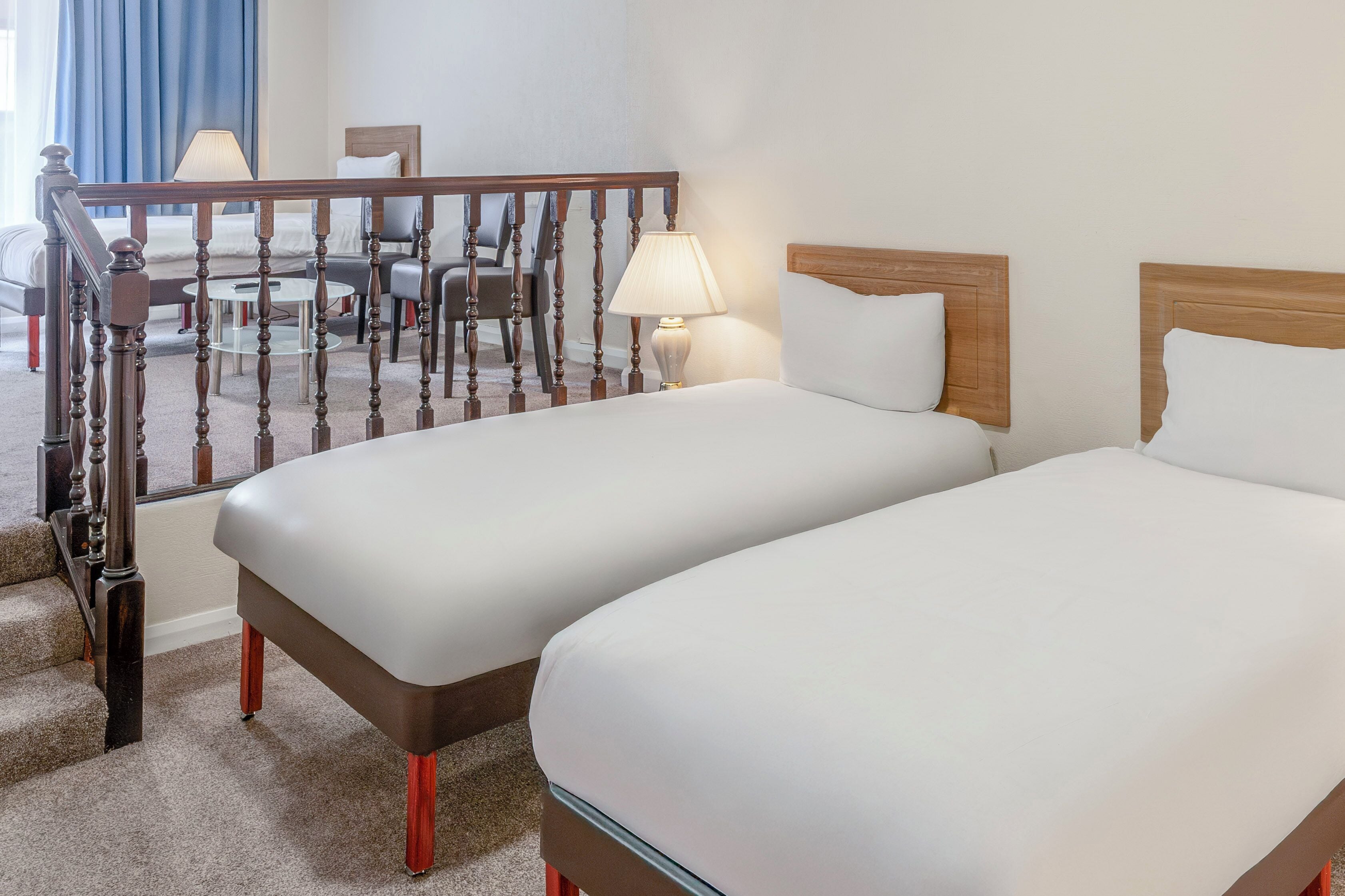 A hotel room with two beds, a wooden staircase, a desk with a chair, and a lamp.