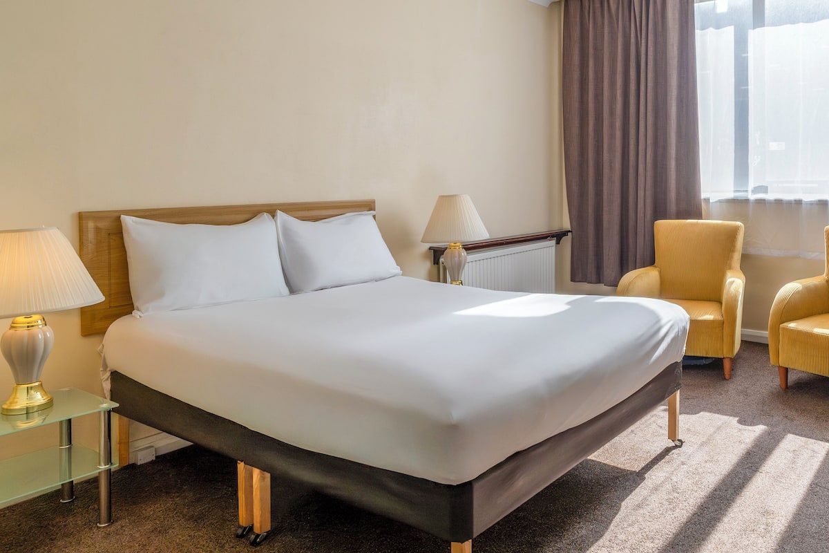 Uma cama bem arrumada com lençóis brancos, uma luminária de cabeceira, uma cadeira e uma janela com cortinas.