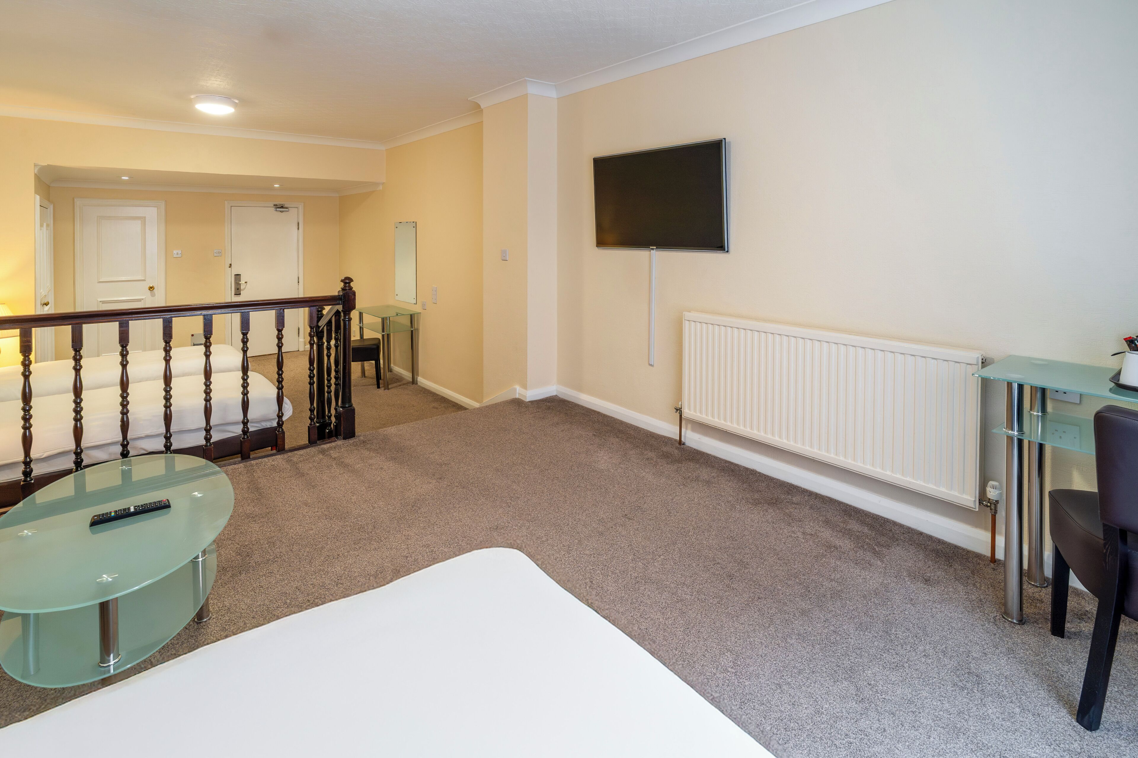 A hotel room with a bed, a glass table with a remote control, a TV mounted on the wall, and a radiator.