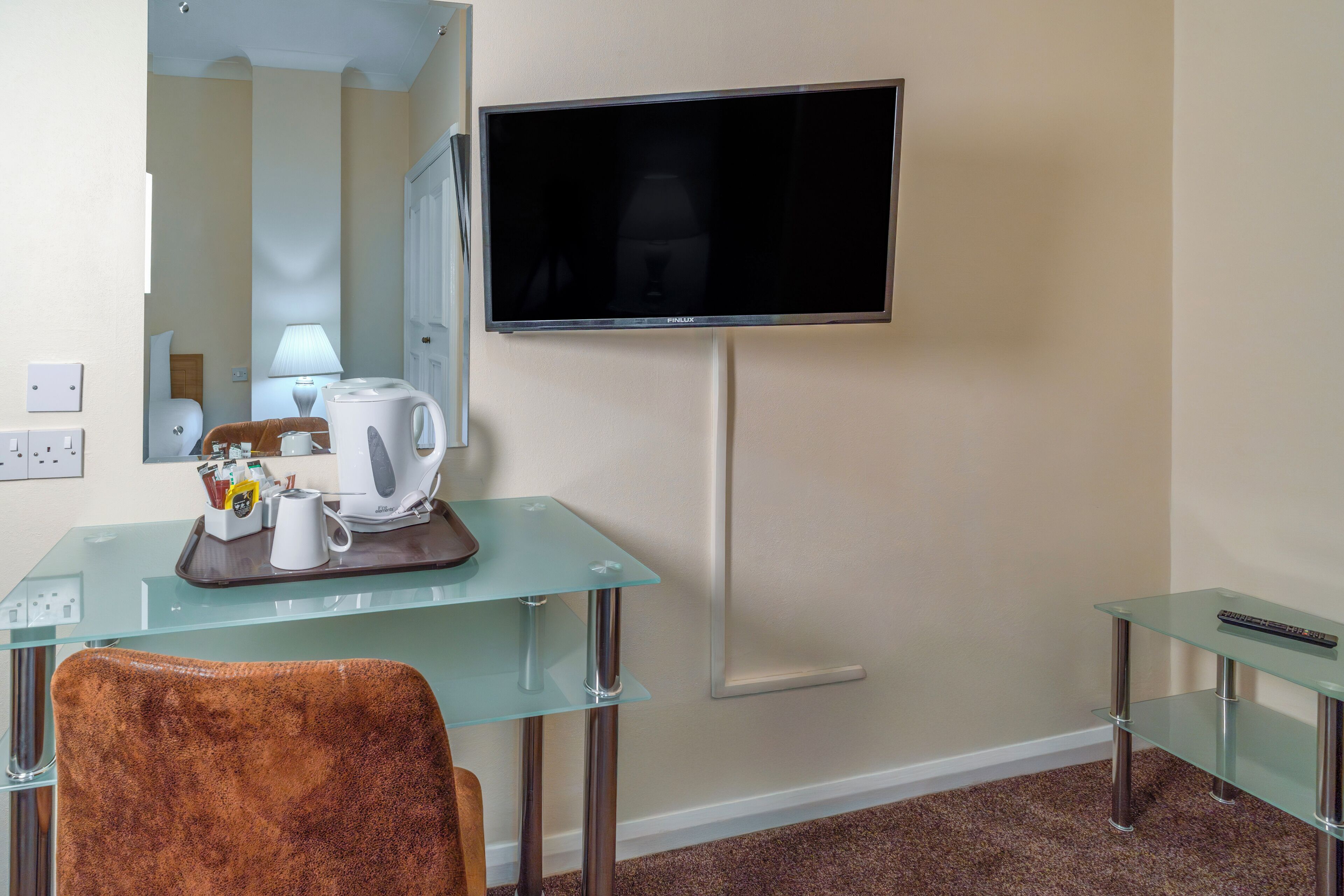 A hotel room with a glass desk, a brown chair, a flat-screen TV mounted on the wall, and a small side table.