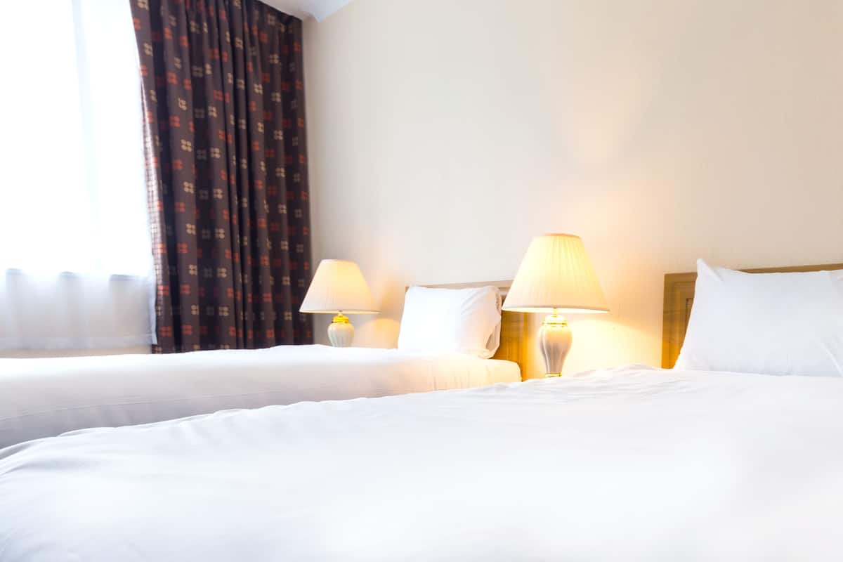Quarto de hotel com duas camas, roupa de cama branca e duas luminárias de cabeceira.