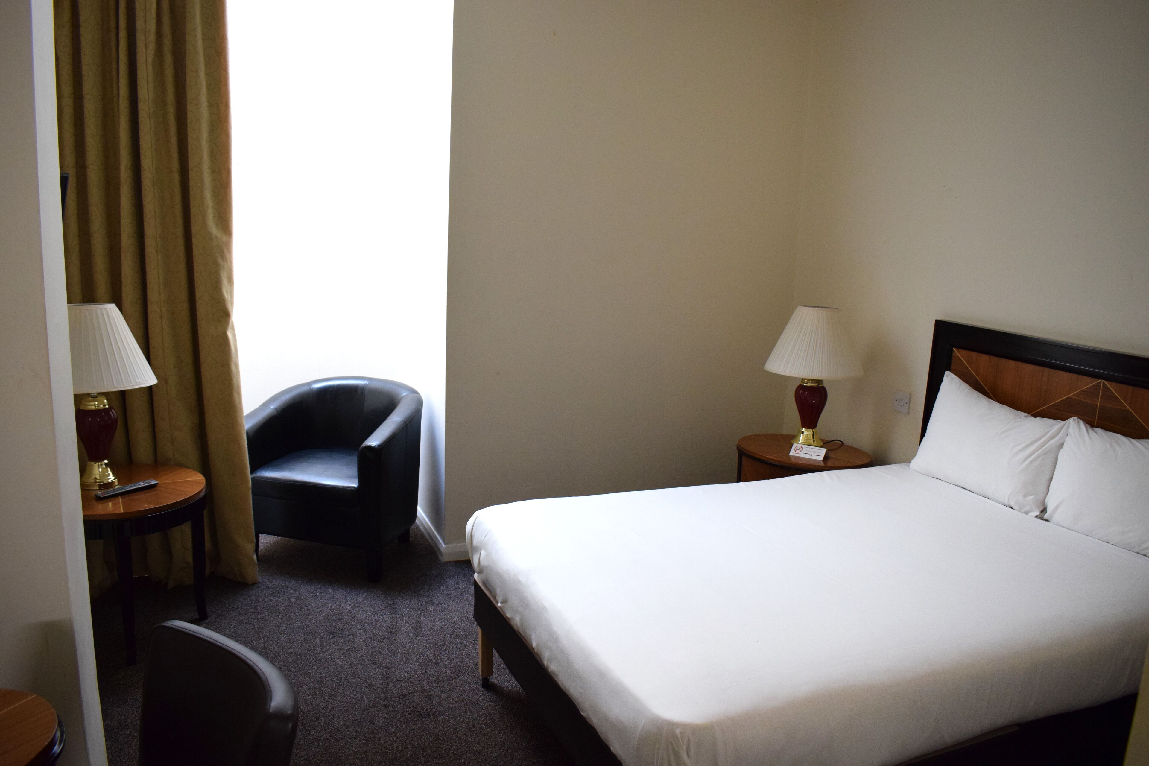Une chambre d’hôtel avec un lit, deux tables de chevet, une chaise, une lampe et une fenêtre avec des rideaux.