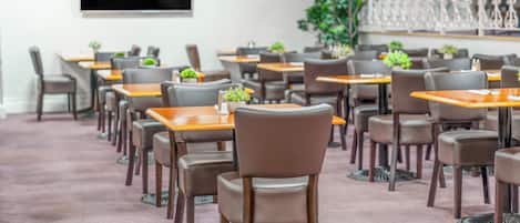 A hotel dining area with multiple tables and chairs, a flat-screen TV mounted on the wall, and decorative plants.