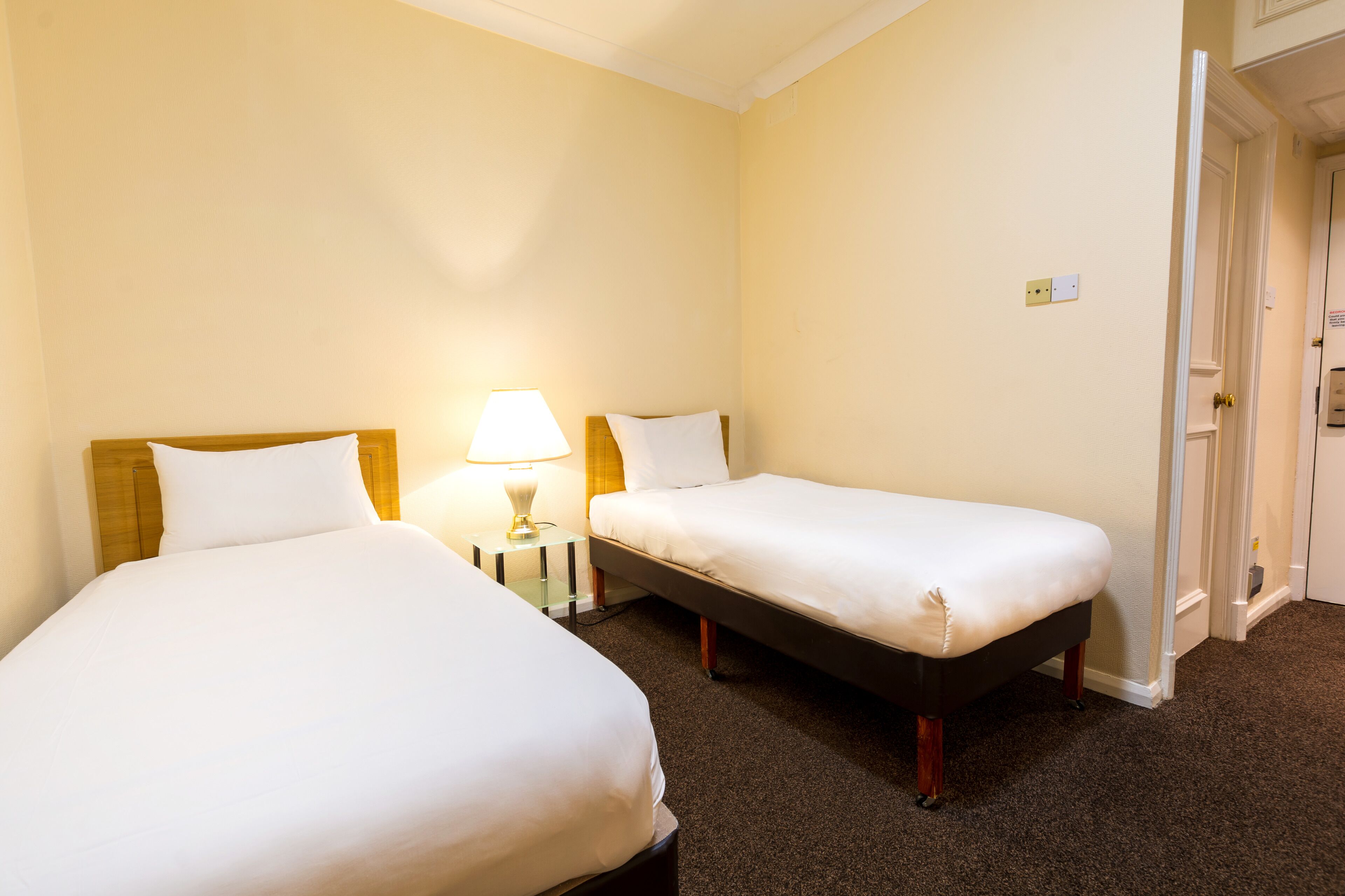 A hotel room with two single beds, a nightstand with a lamp, and a door leading to another room.