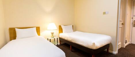 A hotel room with two single beds, a nightstand with a lamp, and a door leading to another room.