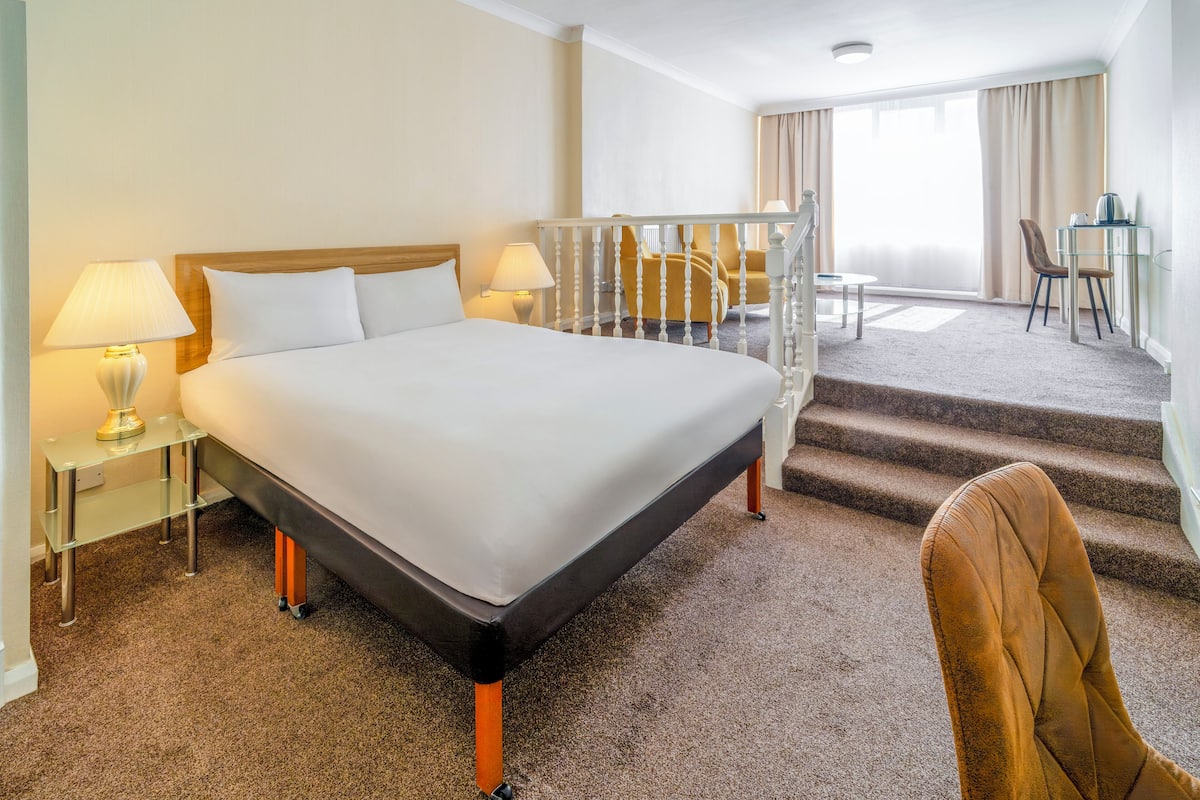 Quarto de hotel com uma cama grande, uma escrivaninha, uma cadeira e uma escada para um andar superior.