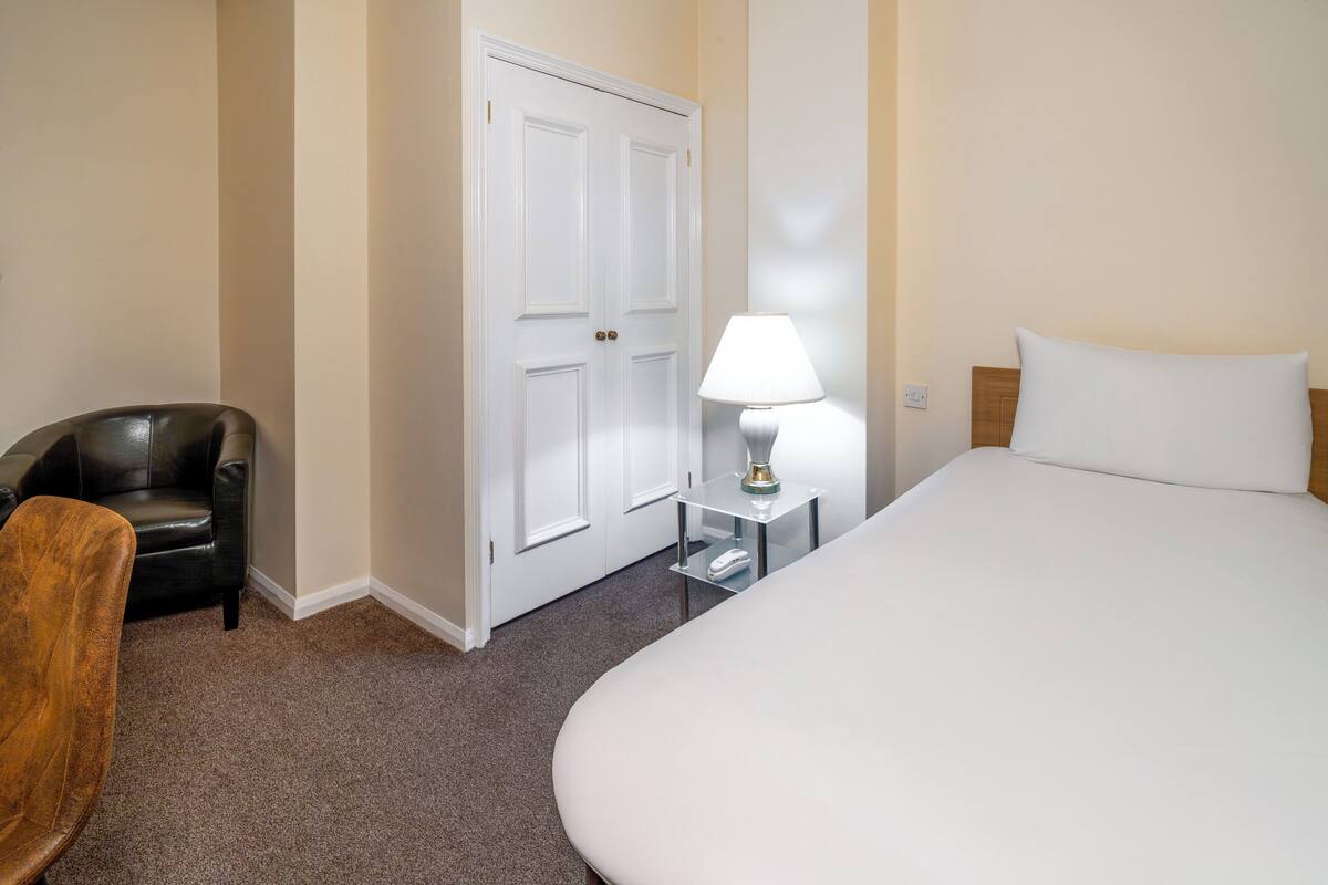 Quarto de hotel com cama, uma cadeira, abajur e guarda-roupa.
