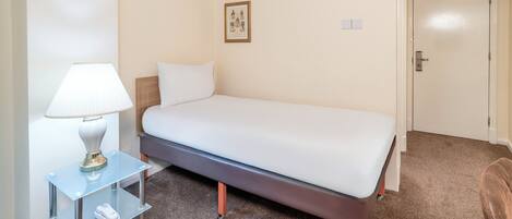 A neatly arranged hotel room with a single bed, a bedside table with a lamp, and a small shelf.