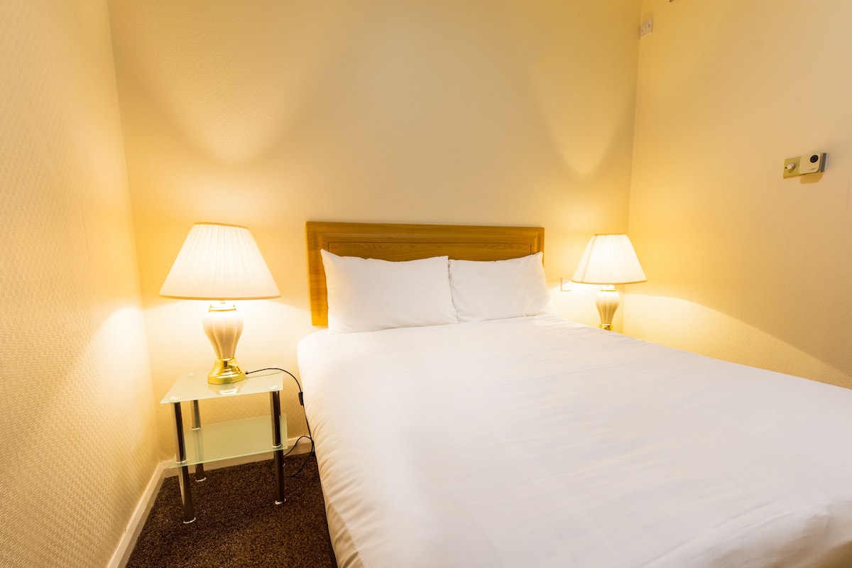 Quarto de hotel com uma cama, duas luminárias de cabeceira e uma mesinha.