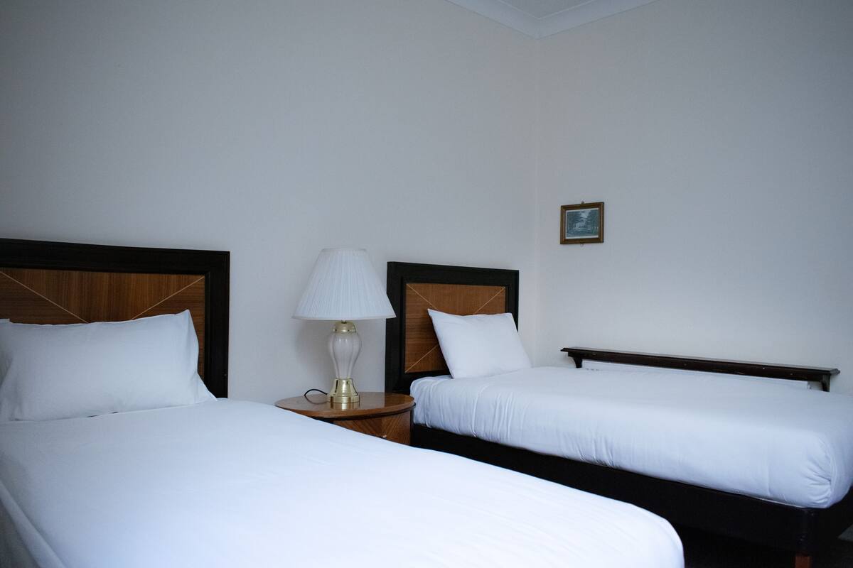 Quarto de hotel com duas camas de solteiro, uma mesa de cabeceira com abajur e uma pequena imagem na parede.