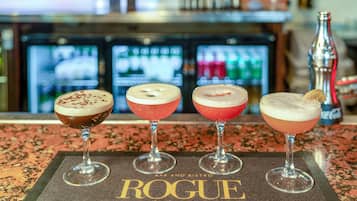 Four cocktails on a bar mat labeled "ROGUE" in a bar setting with a refrigerator and bottles in the background.