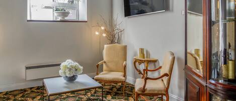 A room with patterned carpet, two armchairs, a small table with a vase, a floor lamp, and a television on the wall.