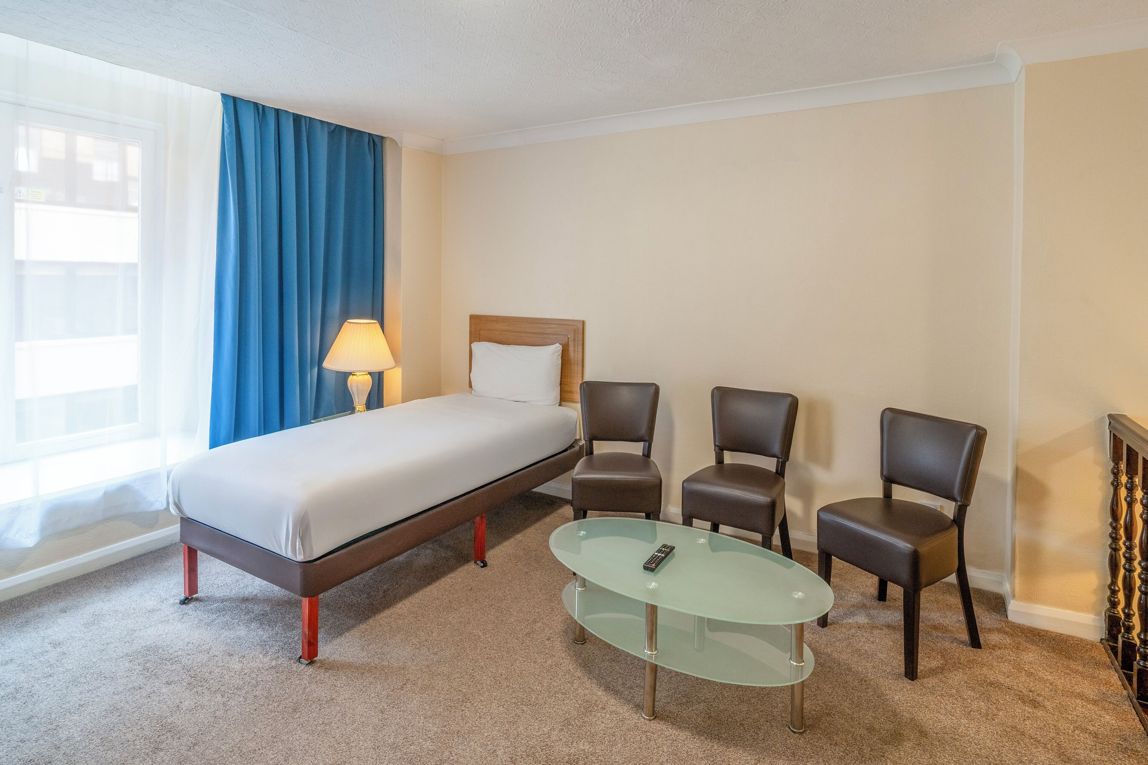 Hotellihuone, jossa on sänky, kaksi tuolia, lasipöytä, lamppu ja ikkuna, jossa on siniset verhot.