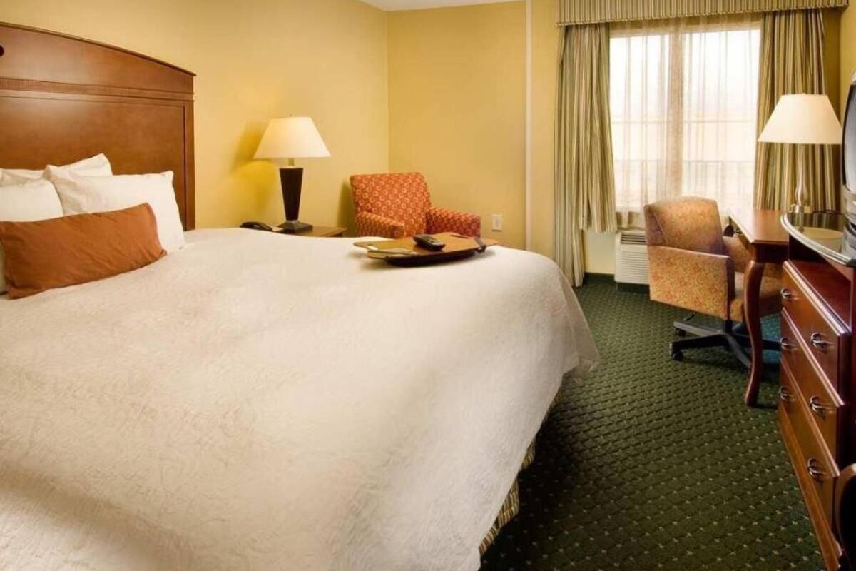 Une chambre d’hôtel avec un grand lit, deux chaises, une commode et une fenêtre avec des rideaux.
