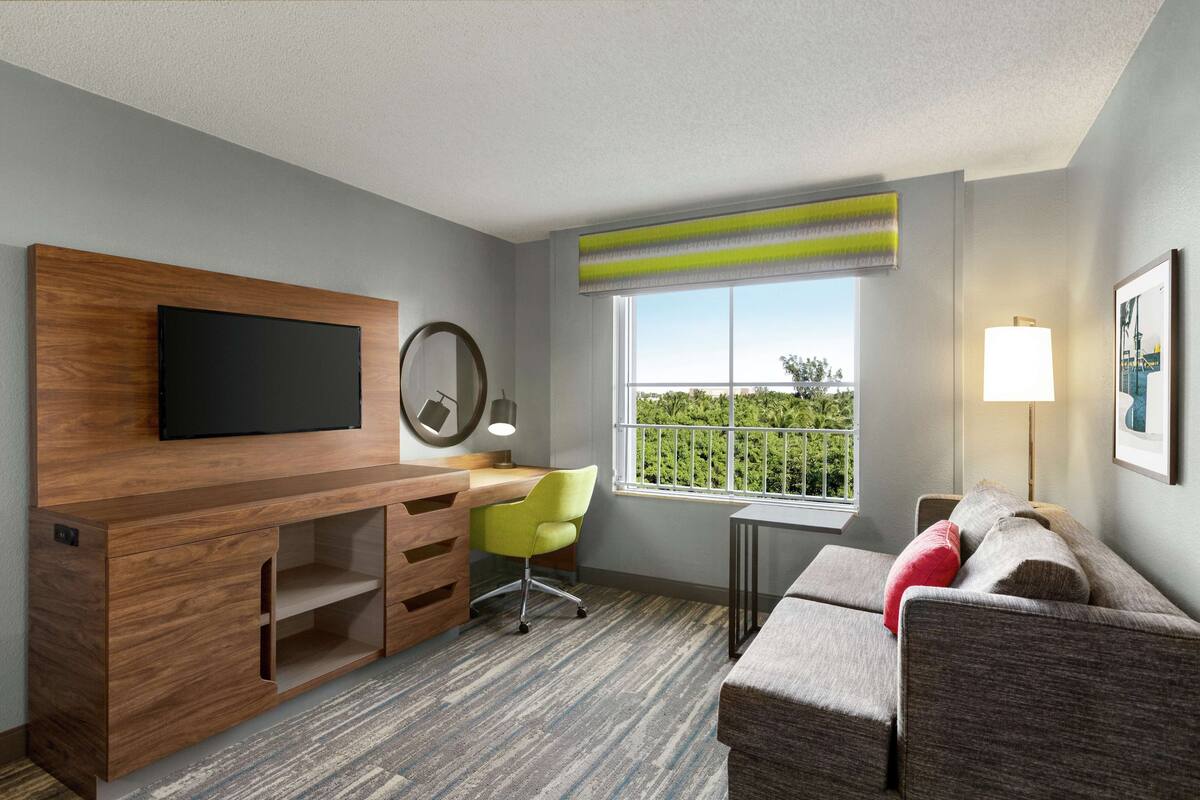 Une chambre d’hôtel moderne avec un bureau en bois, une chaise verte, un canapé gris, un téléviseur fixé au mur, une fenêtre donnant sur un paysage verdoyant et une lampe.