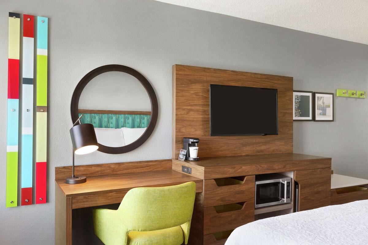Une chambre d’hôtel avec un bureau en bois, une chaise verte, un miroir rond, un téléviseur à écran plat, un four à micro-ondes et un lit avec une literie blanche.