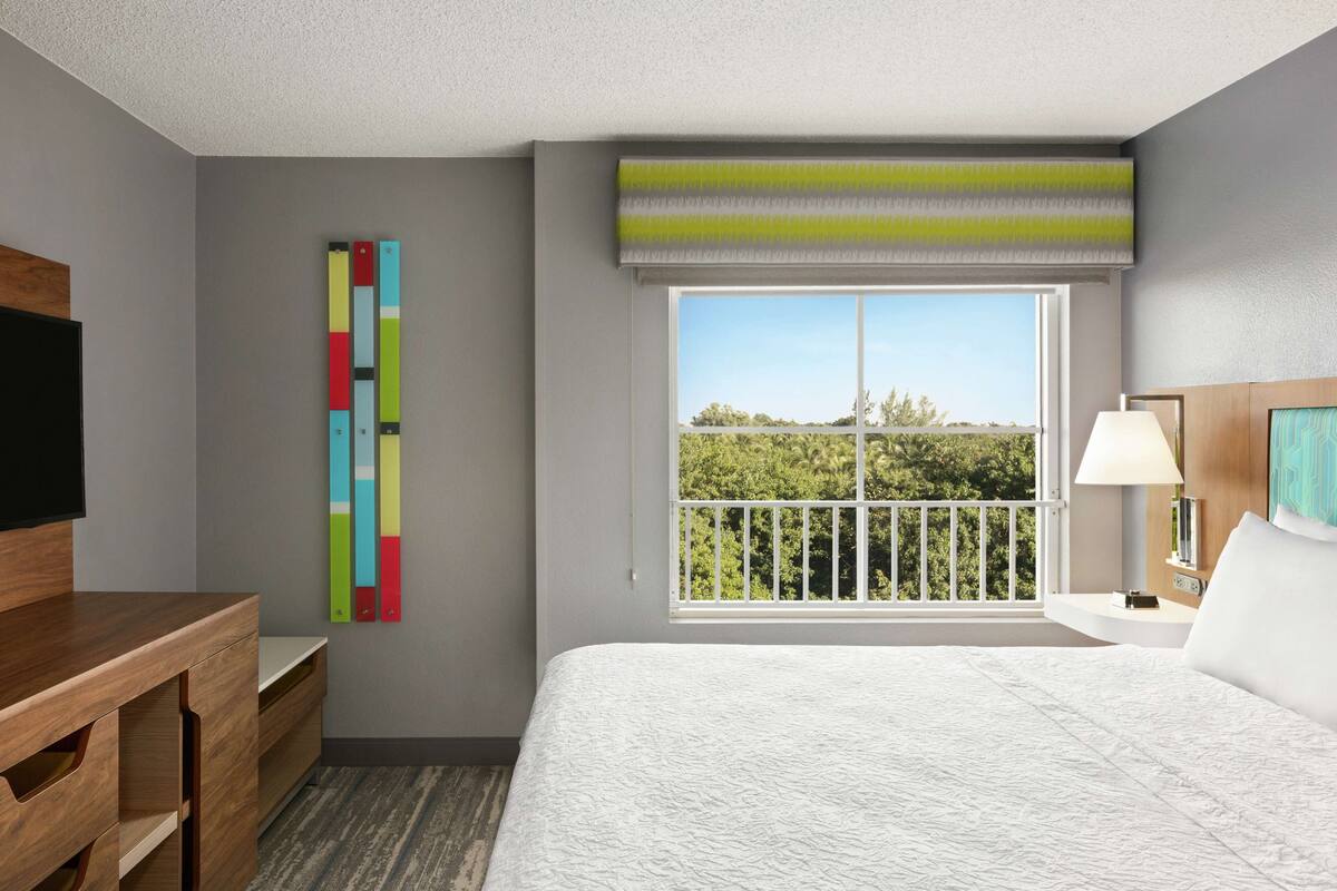 Une chambre d’hôtel avec un lit, un bureau, une télévision, une fenêtre donnant sur un beau paysage et une œuvre d’art colorée accrochée au mur.