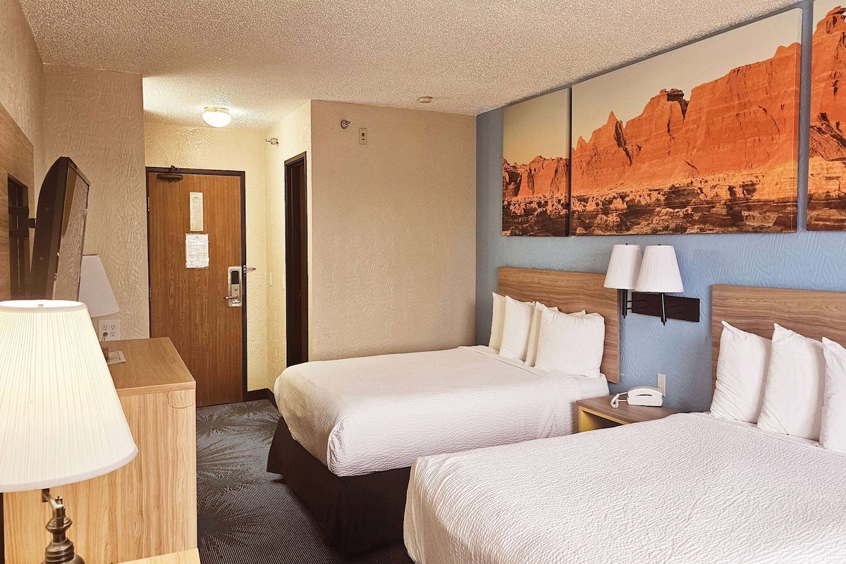 Ein Hotelzimmer mit zwei Betten, einem Holzschrank, einem Schreibtisch mit Lampe und einem großen Wandbild einer Berglandschaft.