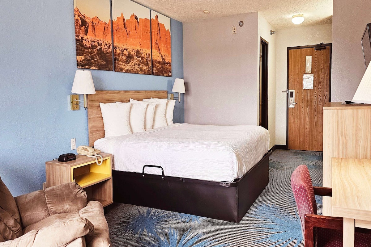 Ein Hotelzimmer mit Bett, Nachttischlampen, einem Holznachttisch, einem Schreibtisch und einem Stuhl.