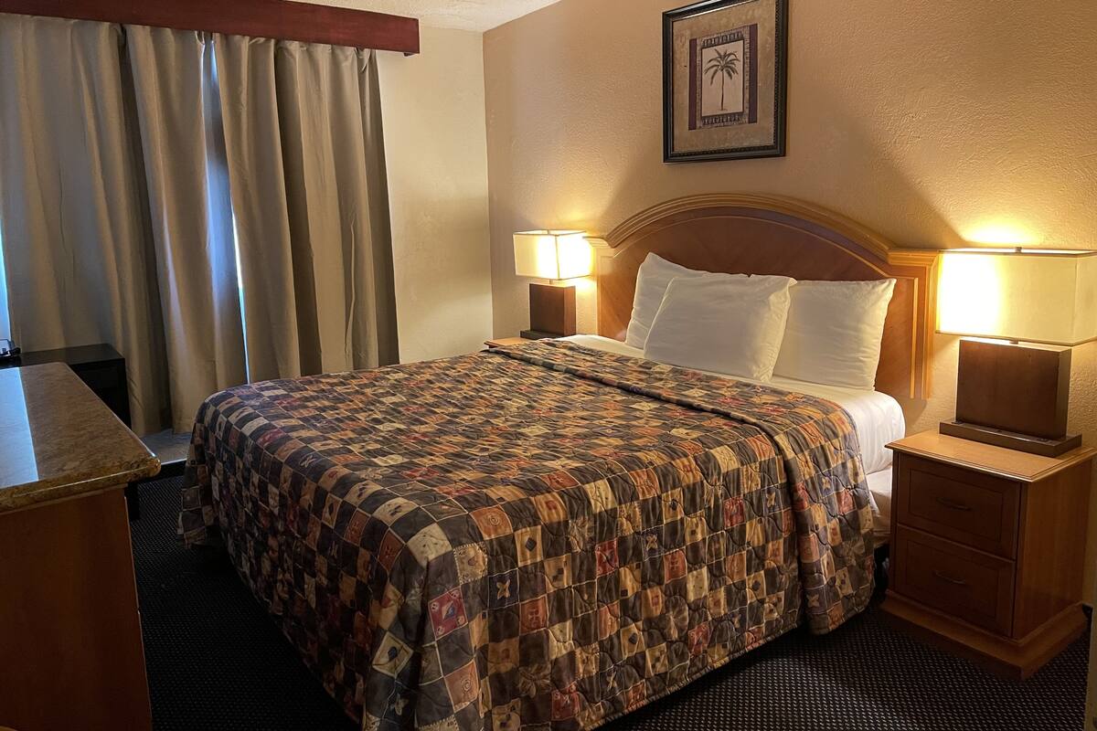A hotel room with a large bed, two bedside lamps, a headboard, a desk, and a window with curtains.