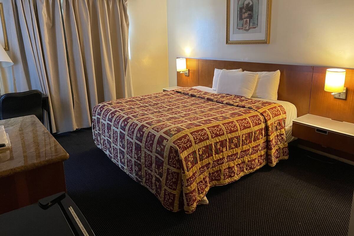 A hotel room with a large bed, a desk, a chair, and a wall-mounted lamp.