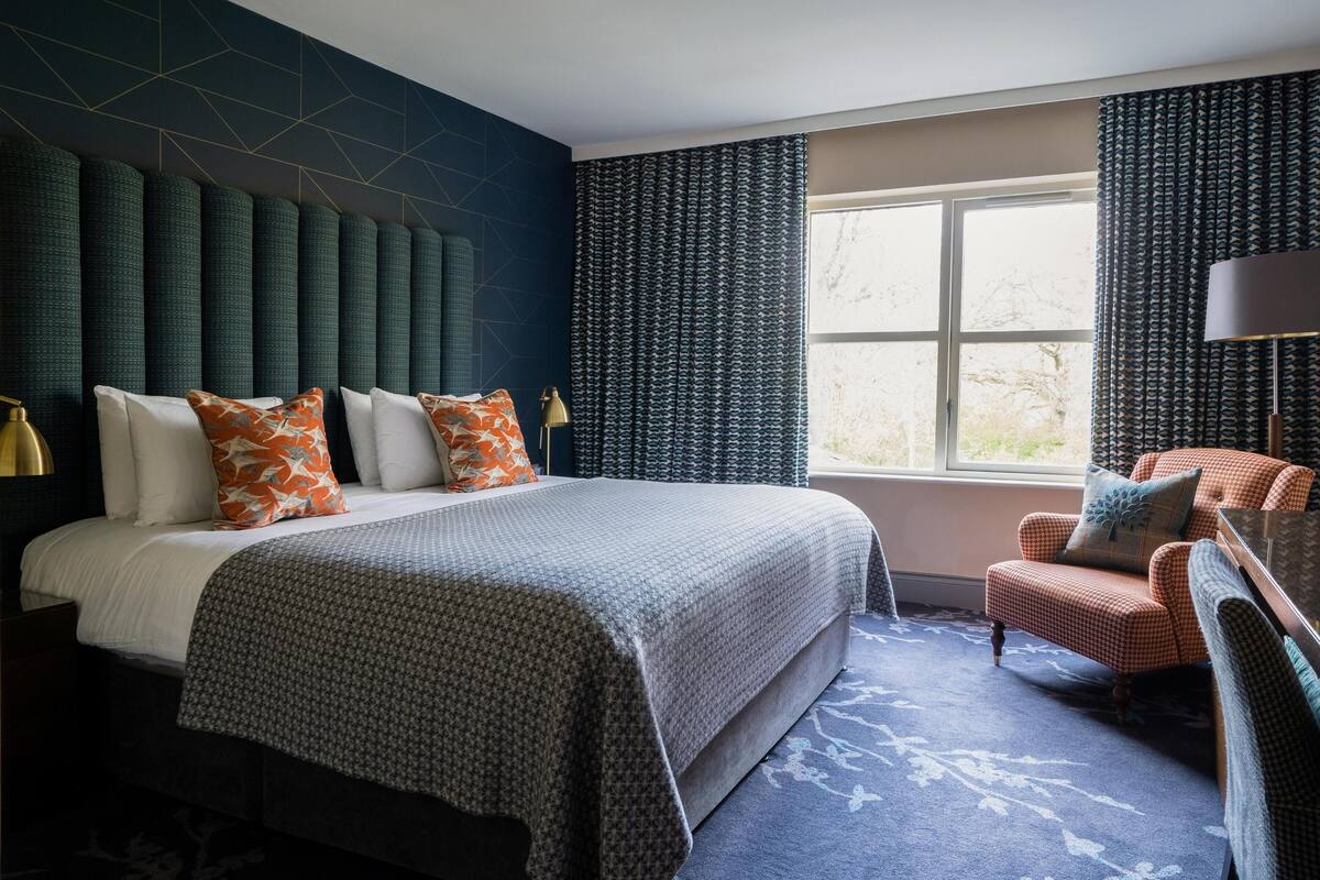 A hotel room with a large bed, a desk with a chair, a window with curtains, and a patterned wall.