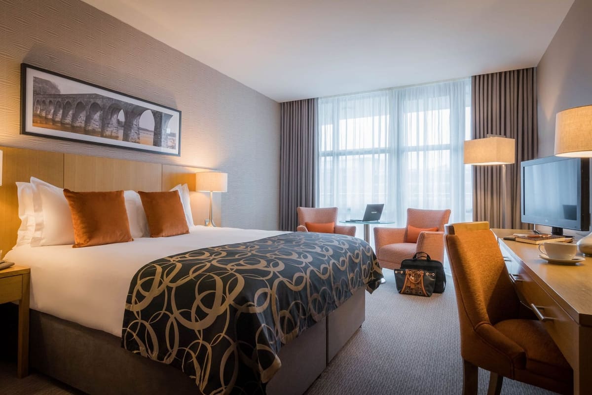 A hotel room with a large bed, two bedside tables, a desk with a chair, a TV, and a window with curtains.