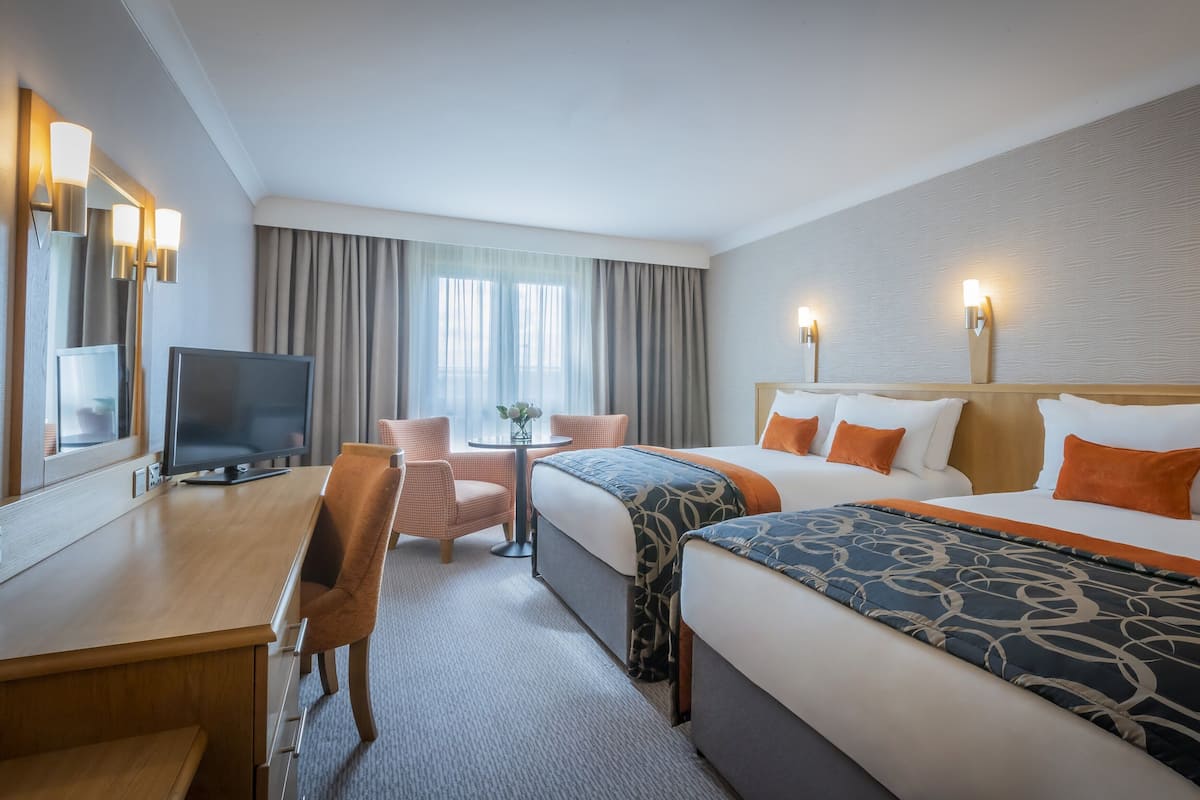 A hotel room with two beds, a desk with a TV, a chair, a small table with a vase, and a window with curtains.