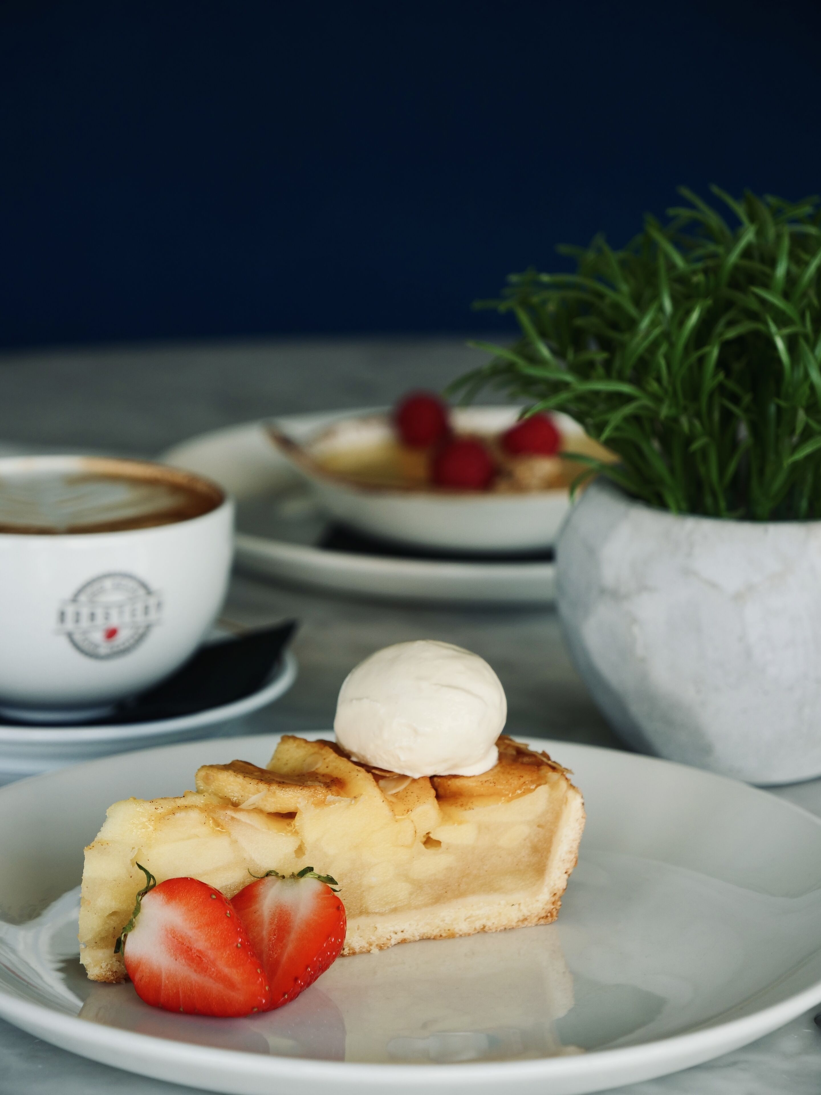 Een nagerechtenbord met een plakje taart, belegd met een bolletje ijs en een aardbei, een kop koffie en een schaal fruit.