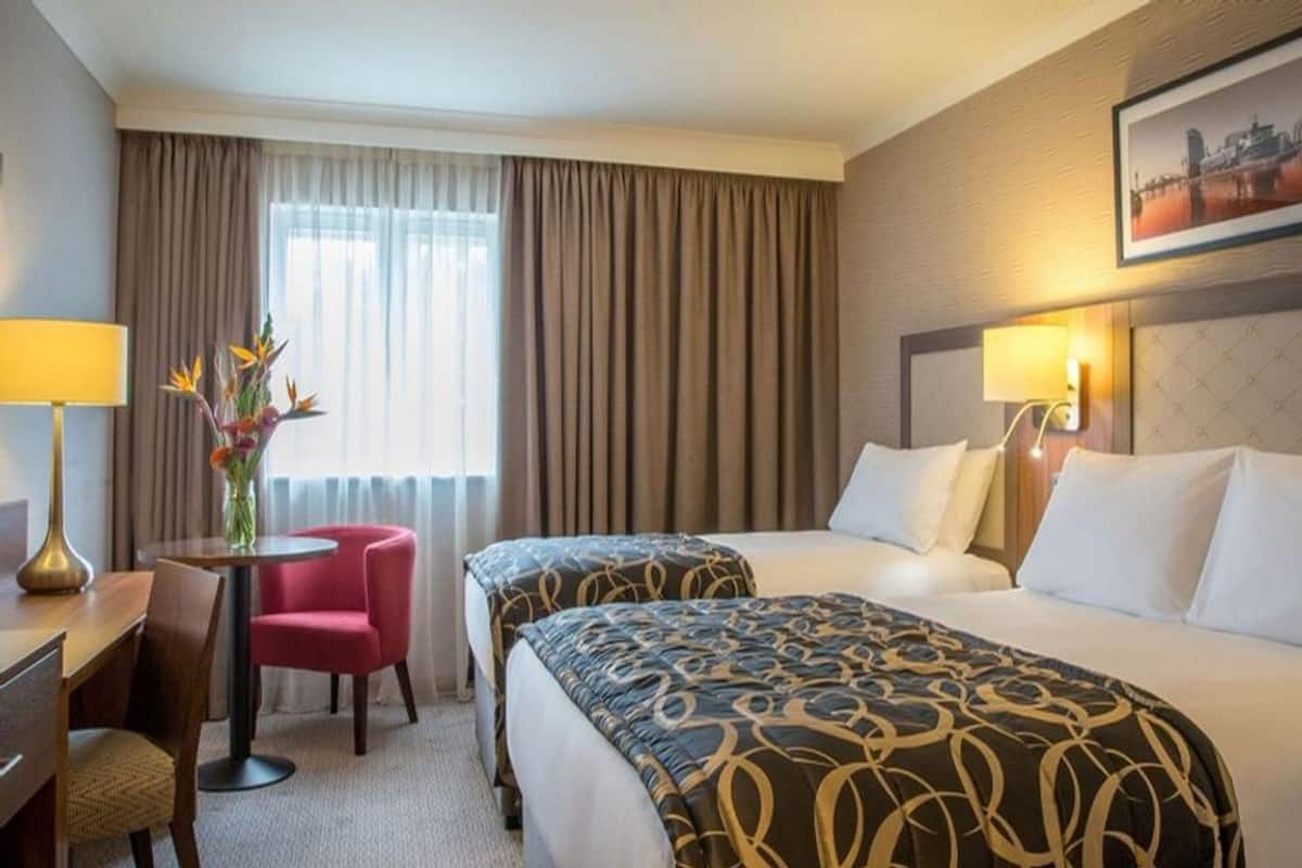 A hotel room with two beds, a desk, a chair, a vase with flowers, and a cityscape painting on the wall.
