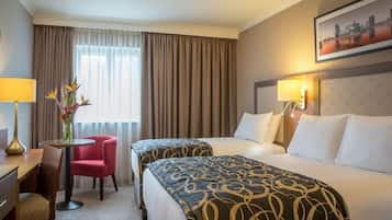 Habitación de hotel con dos camas, un escritorio, una silla, un jarrón con flores y un cuadro de la ciudad en la pared.