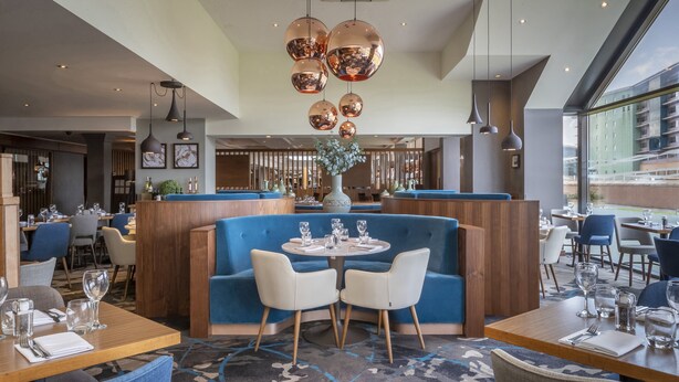 Et moderne restaurantinteriør med et centralt siddeområde, der inkluderer en blå bænk, hvide stole og et rundt bord.
