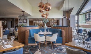 A modern restaurant interior with a central seating area featuring a blue booth, white chairs, and a round table. - Clayton Hotel, Manchester Airport (Manchester)