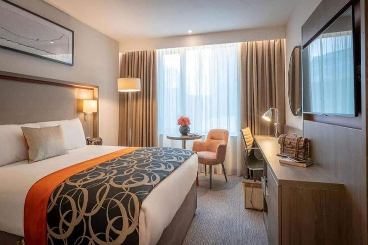 A hotel room with a large bed, a desk with a chair, a small table with a vase, and a TV.