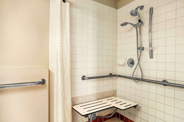 A bathroom with white tiled walls, a shower with a handheld showerhead and a handheld faucet, a grab bar, and a bench.