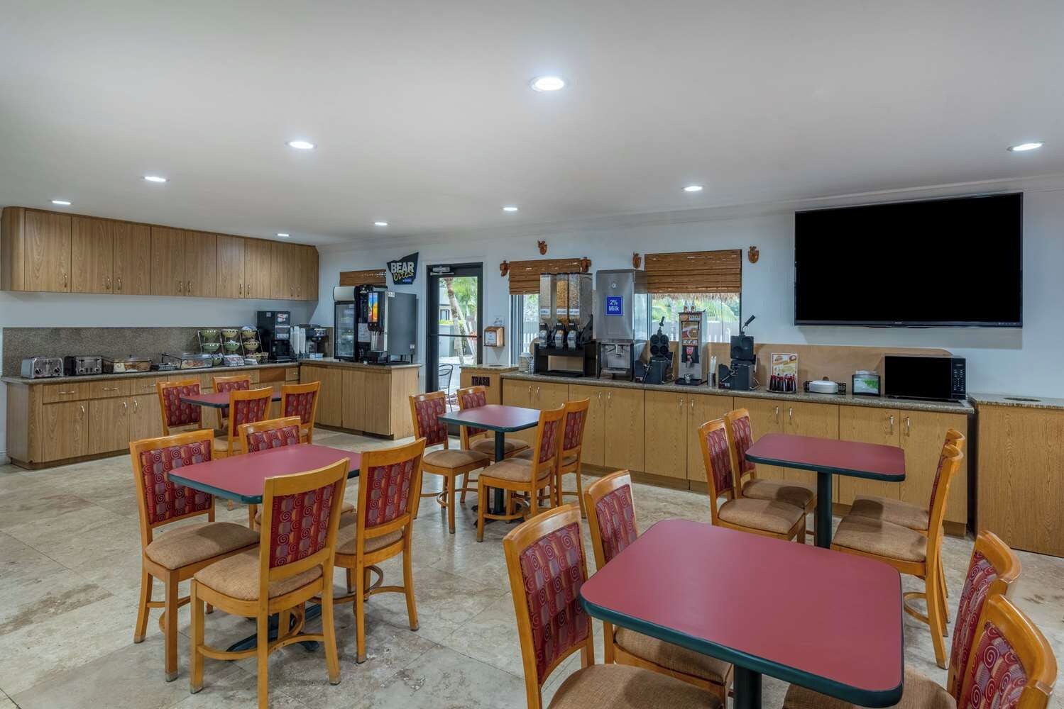 Un coin repas avec des tables et des chaises en bois, une cuisine équipée d’appareils électroménagers et un téléviseur à écran plat fixé au mur.