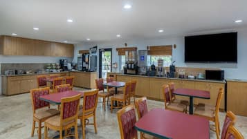 A dining area with wooden tables and chairs, a kitchen with appliances, and a flat-screen TV on the wall.