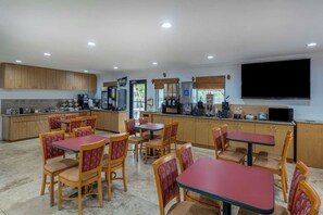 Un coin repas avec des tables et des chaises en bois, une cuisine équipée d’appareils électroménagers et un téléviseur à écran plat fixé au mur.