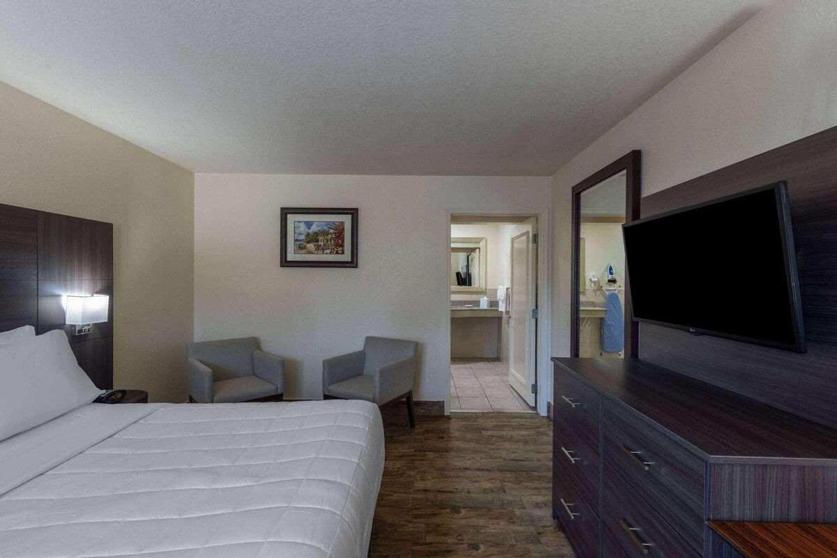 A hotel room with a bed, two armchairs, a desk with a flat-screen TV, a dresser, and a bathroom visible through an open door.