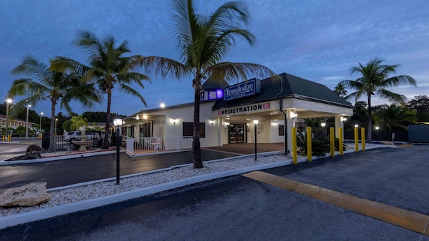 Un edificio Travelodge con una mesa de recepción, palmeras y un estacionamiento al anochecer.
