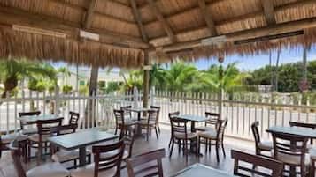 An outdoor patio with tables and chairs, a thatched roof, and a view of palm trees.