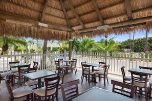 Une terrasse extérieure avec des tables et des chaises, un toit de chaume et une vue sur des palmiers.