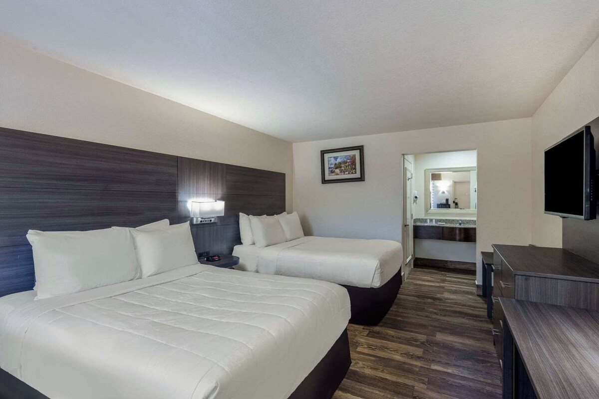 A hotel room with two beds, a TV, a desk, a mirror, and a bathroom visible through an open door.