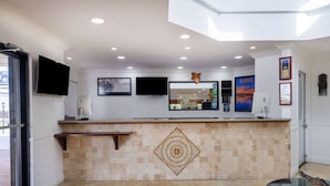Zona de recepción de un hotel con suelo de baldosas, mostrador de recepción, televisor montado, obras de arte enmarcadas y un elemento decorativo en la pared.