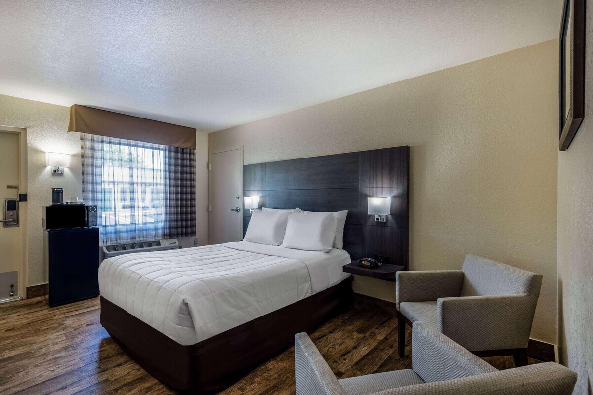 A hotel room with a bed, bedside table, chair, and a window with curtains.